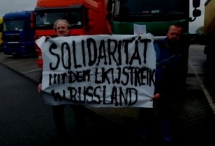 Two truck drivers standing in a truck parking area holding a banner with the slogan “Solidarity with the truck driver's strike in Russia”